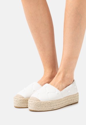 Gambe che indossano scarpe espadrillas bianche in soffice tessuto imbottito con suole in giuta intrecciata, mostrate su uno sfondo chiaro e semplice.