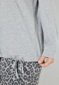 Persona che indossa un top a maniche lunghe grigio chiaro con orlo regolabile e pantaloni grigi con stampa leopardata, mentre si tiene da un lato del top.