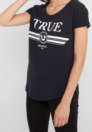 Kobieta ubrana w czarną koszulkę True Religion z białym logo i paskami, dobraną do czarnych spodni na jednolitym tle.