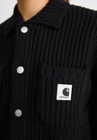 Schwarze gestrickte Strickjacke mit gerippter Textur, kragenförmigem Design, silbernen Druckknöpfen und einem weißen Etikett mit Logo auf der Brusttasche.