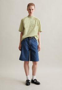 Maglietta verde chiaro a maniche corte con colletto rialzato, abbinata a pantaloni corti in denim blu. Indossata con scarpe nere e calze bianche. Design semplice.