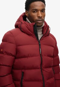 Homme portant une veste matelassée rouge avec capuche et poches zippées, superposée sur un pull col roulé beige.