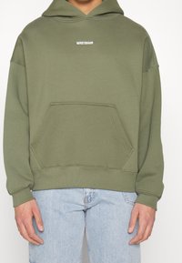 Personne portant un sweat à capuche vert olive avec le texte "WRSTBHVR" sur la poitrine et un jean bleu clair, debout devant un fond blanc.