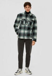 Jachetă din flanel în carouri negre, albe și teal, cu două buzunare pe piept, închidere cu nasturi și o croială ușor supradimensionată. Pantaloni negri și adidași.