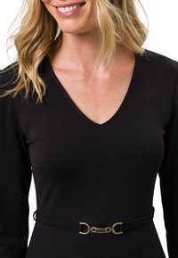Femme blonde souriante, portant un haut noir à manches longues et col en V avec une fine ceinture noire équipée d'une petite boucle dorée.