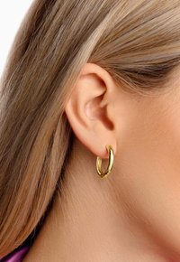 Goldene Creolen mit hochglänzender Oberfläche, die ein gedrehtes Design aufweisen und eine mittlere Größe haben, getragen im Ohr eines Models.