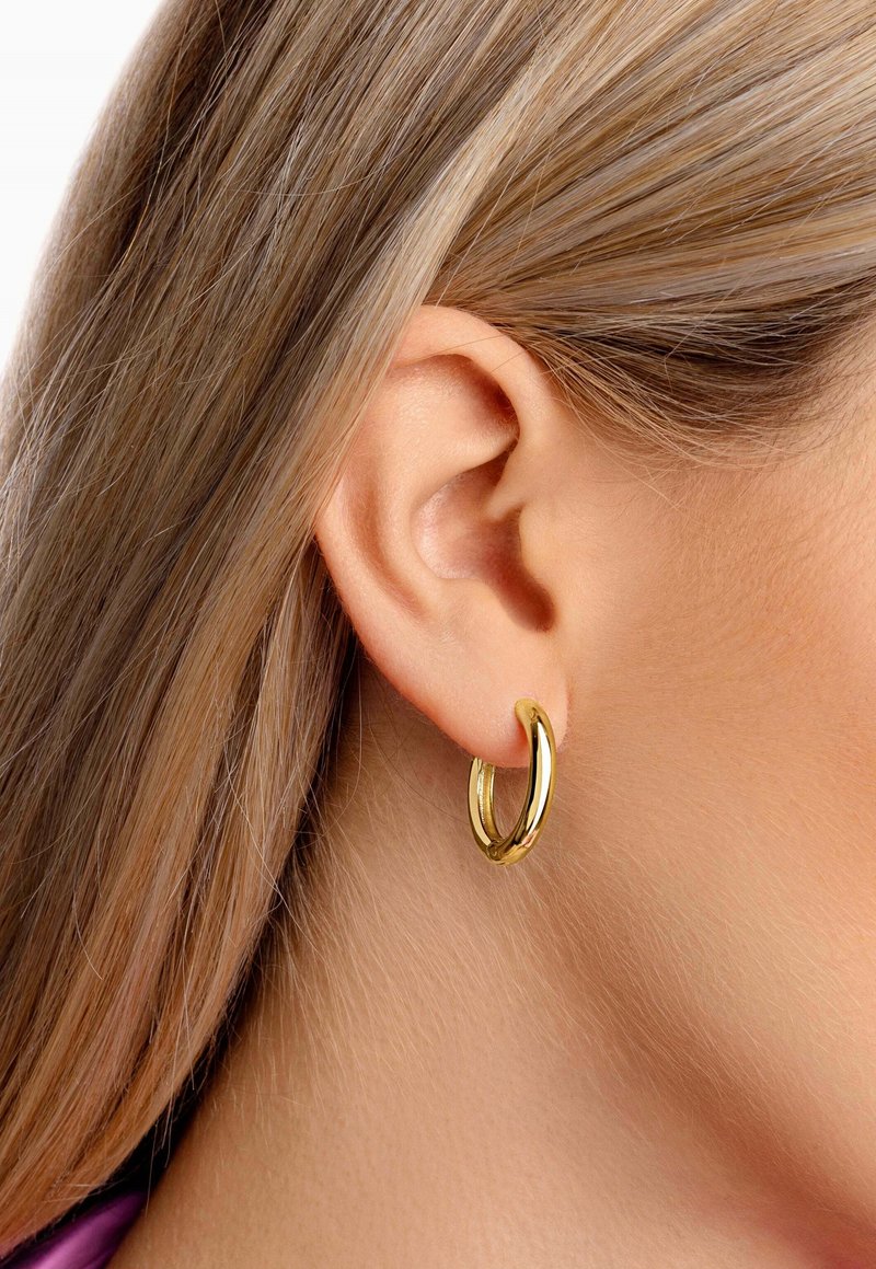 Goldene Creolen mit hochglänzender Oberfläche, die ein gedrehtes Design aufweisen und eine mittlere Größe haben, getragen im Ohr eines Models.