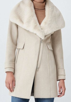Beżowy wełniany płaszcz z dużym kołnierzem z sztucznego futra, asymetrycznym suwakiem, kieszeniami z klapkami oraz mankietowanymi rękawami, noszony na beżowym golfie.