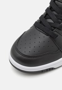 Schwarzer Sneaker mit einem strukturierten Leder-Obermaterial, Perforationen an der Zehenpartie, schwarzen Schnürsenkeln und einer weißen Gummisohle mit schwarzen Akzenten.