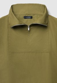 Pullover verde oliva con colletto alto, chiusura anteriore con zip e tasca rettangolare sul petto. Presenta una texture liscia e opaca. Etichetta del marchio visibile.