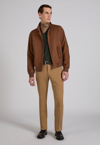 Uomo con giacca marrone con zip, camicia verde scuro, sciarpa fantasia al collo, pantaloni color camoscio, cintura scura e scarpe nere, in piedi davanti a uno sfondo grigio chiaro.