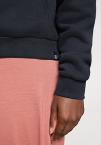 O mână închisă la culoare, relaxată lângă corp, îmbrăcată cu un pulover negru cu mâneci lungi și pantaloni roz, pe un fundal simplu deschis.