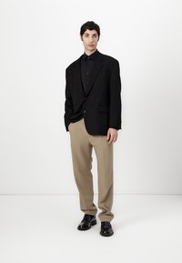 Crni oversized sako preko crne košulje na dugmad, u kombinaciji s bež hlačama i crnim kožnim cipelama. Glatka tkanina, krojen dizajn.