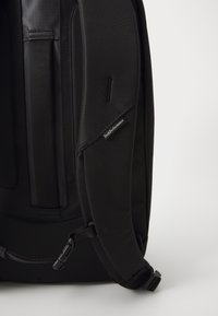 Svart ryggsekk laget av teksturert nylon, med polstrede skulderstropper, en logoetikett og et slankt, moderne design med justerbare detaljer.