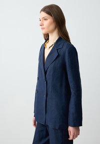 Młoda kobieta w granatowym blezrze i spodniach, nosząca naszyjnik z wielokolorowych koralików, stojąca na jednolitym jasnoszarym tle.