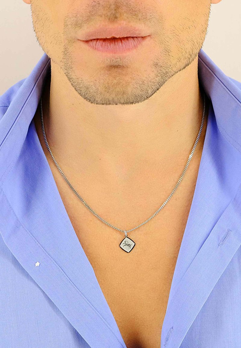 Collier en chaîne argentée avec un pendentif en forme de losange orné d'un motif gravé, porté par-dessus une chemise à col bleu clair.