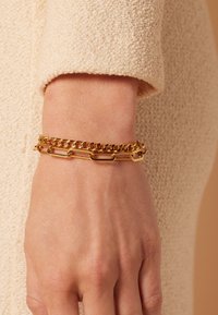 Gouden metalen armband met in elkaar verkerende schakels en platte rechthoekige accenten, gedragen om een pols tegen een gestructureerde, neutrale achtergrond.