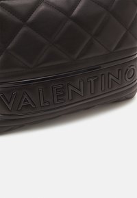 Prošívaná černá kožená kabelka s vyvýšeným pruhem loga Valentino napříč dolní částí na bílém pozadí.