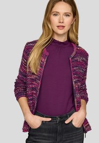 Mehrfarbige lila Strickjacke mit strukturierter Oberfläche, Frontreißverschluss und gerippten Bündchen, getragen über einem einfarbigen lila Oberteil.