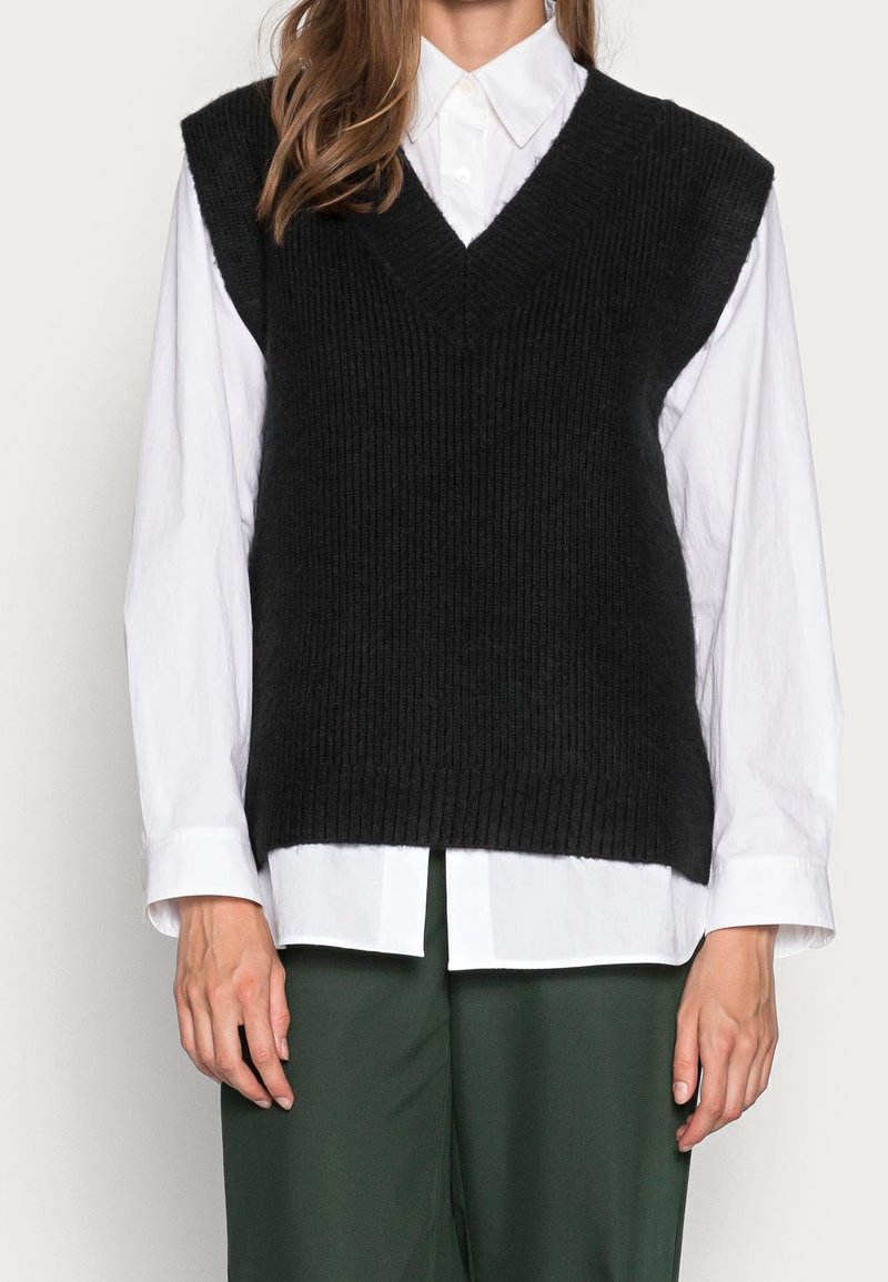 Femme portant un gilet sans manches en maille noire sur une chemise blanche boutonnée et un pantalon vert foncé, debout devant un fond uni.