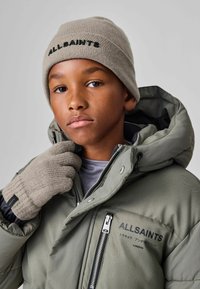 Zaļš pūkains džemperis ar kapuci, ar uzrakstu "ALLSAINTS". Bēšs adīts beanie un cimdi, ar etiķeti. Gluda tekstūra, funkcionāls dizains.