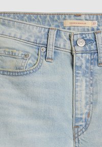 Jean en denim bleu avec un design évasé, délavage clair, couture visible et une poche avant. Comprend un bouton en métal et un fermoir.