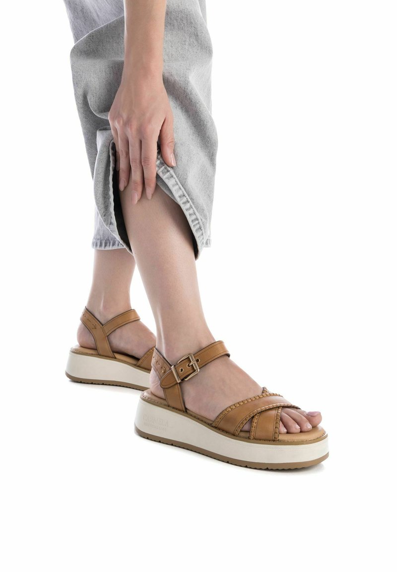 Sandalias de cuero marrón con tira en el tobillo, detalles texturizados y suela gruesa blanca. Llevadas con pantalones grises claros, remangados.