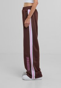 Pantalones deportivos marrones con franjas blancas y rosas a los lados, que presentan la palabra "FELICIOUS" en rosa negrita en la pierna izquierda. Cintura elástica.