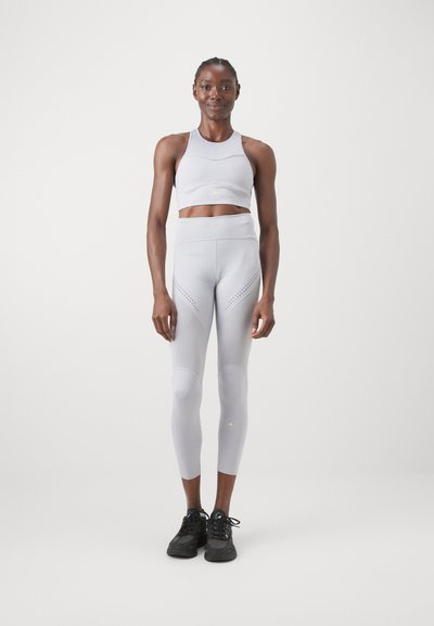 Világosszürke sportmelltartó és leggings szett, lélegző hálós részletekkel, varrás nélküli dizájnnal és magas derékkal. Fekete sportcipők egészítik ki a megjelenést.