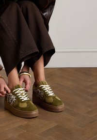 Green suede sneakers with white laces, circular accents, and a textured base. The upper features a patterned detailing on the sides.