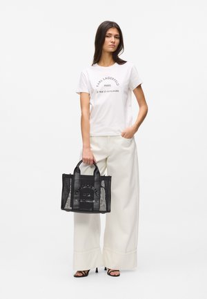 Femme portant un t-shirt blanc Karl Lagerfeld et un pantalon à jambes larges, tenant un sac fourre-tout en mesh noir, debout sur un fond blanc.