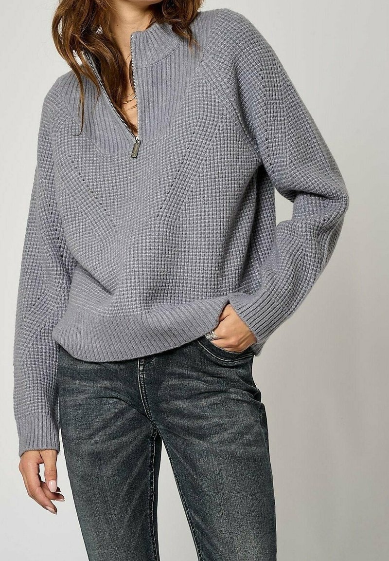 Pull gris en maille avec col à demi-fermeture éclair, motifs texturés et manches côtelées, associé à un jean en denim foncé.