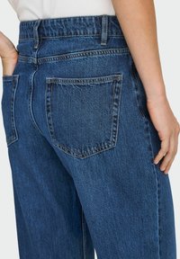 Personne portant un jean en denim bleu avec des poches arrière, une main reposant dans une poche et l'autre main détendue le long du corps.