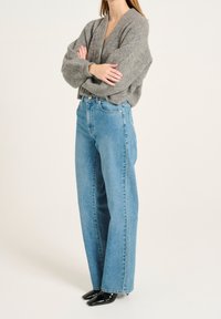 Graue, oversized gestrickte Strickjacke mit V-Ausschnitt und großen Bündchen, kombiniert mit hochgeschnittenen blauen Denim-Jeans. Schwarze spitze Schuhe runden den Look ab.