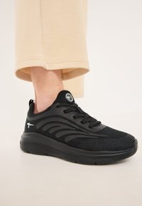 Black athletic shoe with mesh upper, textured accents, TPU support overlays, and a thick sole. Features a logo on the tongue.