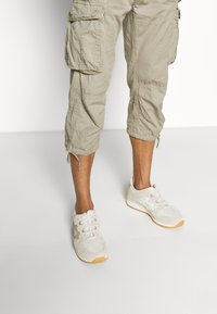 Shorts cargo beige avec plusieurs poches, fabriqués dans un tissu léger, associés à des chaussures de sport blanches avec une semelle en gomme.
