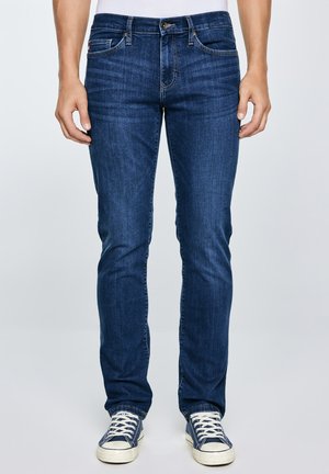 Mees, kes kannab tumedat sinist kitsaid teksapükse ja mereväe siniseid madalaid tossusid valgete paeltega, seisab käed lõdvalt külgedel.