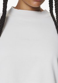 Sudadera blanca con escote acanalado, que presenta un sutil logo en relieve en el pecho. Textura suave, diseño casual, estilo minimalista.