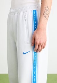 White jogger pants with a blue Nike logo and a side zipper pocket. Features a blue stripe with repeated "NIKE" text along the outer leg.