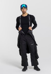 Czarne spodnie narciarskie z regulowanymi ramiączkami, zapinanymi na suwak kieszeniami i wodoodpornym materiałem, noszone na czarnej, długorękawowej warstwie bazowej oraz butach.