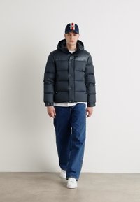 Giacca puffer grigia scura con cappuccio, caratterizzata da un design imbottito. Abbinata a pantaloni in denim blu e sneakers bianche per un look informale.