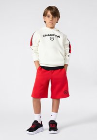Witte hooded sweatshirt met "CHAMPION" tekst, rode en zwarte strepen op de mouwen. Rode shorts, zwarte sportschoenen met rode accenten, witte sokken.
