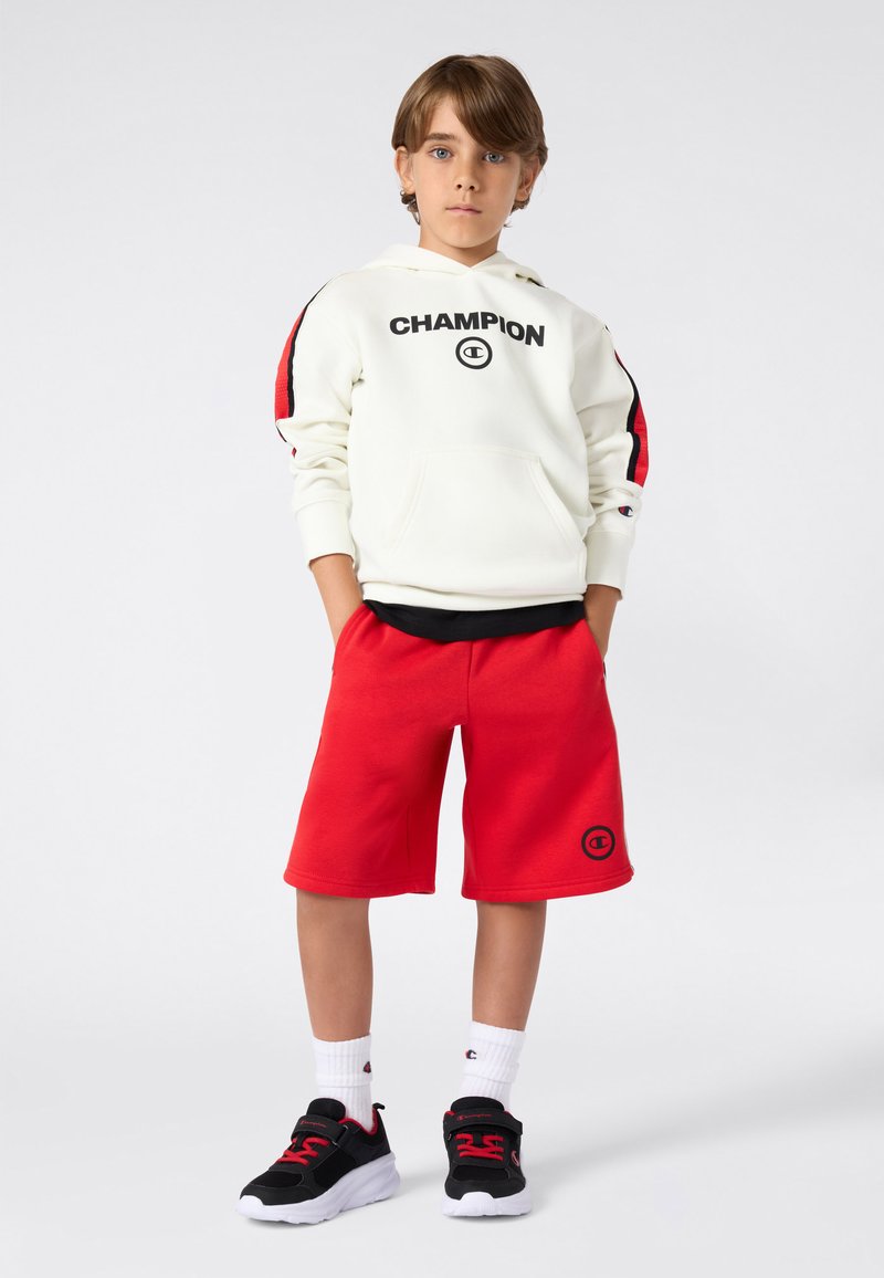 Witte hooded sweatshirt met "CHAMPION" tekst, rode en zwarte strepen op de mouwen. Rode shorts, zwarte sportschoenen met rode accenten, witte sokken.