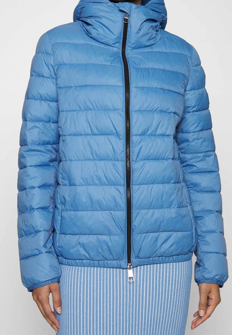 Veste matelassée bleue avec coutures horizontales, fermeture éclair à l'avant, col montant et poignets élastiqués, portée sur une jupe rayée bleu et blanc.