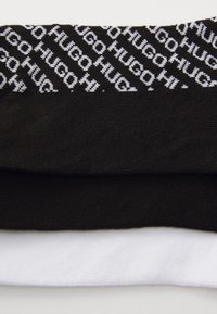 Trois paires de chaussettes : une noire avec un logo "HUGO" répété en blanc, une noire unie et une blanche unie, le tout en coton doux.
