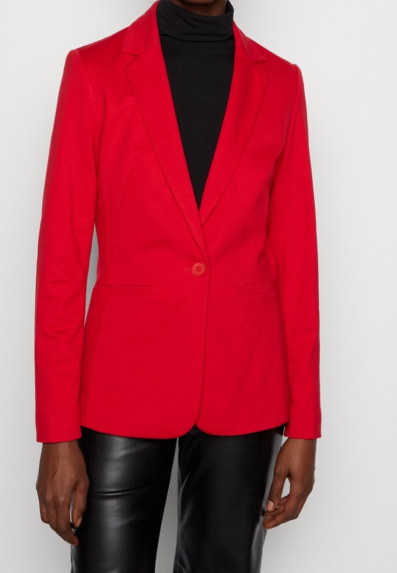 Czerwona marynarka z wyłogami i zapięciem na jeden guzik. Posiada dopasowany krój i gładki materiał, połączona z czarnym golfem.