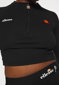 Czarny krótki top z zamkiem błyskawicznym przy dekolcie, z haftowanymi logo Ellesse i kolorowym emblematem na piersi. Miękki, elastyczny materiał.