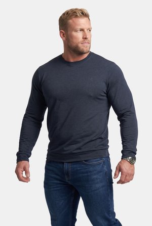 Mann mit blondem Haar, der ein dunkelblaues Langarmhemd und blaue Jeans trägt, schaut zur Seite mit einer silbernen Armbanduhr am linken Handgelenk.