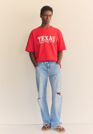 Junger Mann trägt ein rotes Texas Copper Canyon T-Shirt, zerrissene hellblaue Jeans und braune Sandalen und steht mit den Händen in den Taschen.