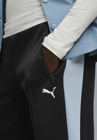 Zwarte en lichtblauwe sportbroek met een ritszak en een wit Puma-logo aan de zijkant. De stof lijkt textuur te hebben en sportief te zijn.